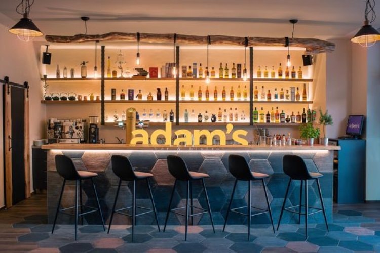 Stylový bar Adam’s s podsvíceným nápisem, vysokými barovými židlemi a dekorativním osvětlením.